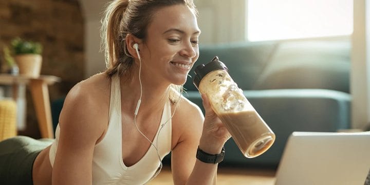 A woman smiling in front of her laptop while drinking protein shake
