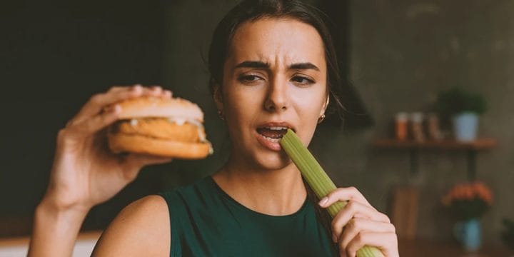 a woman holding a burger with one hand and eating a vegetable with the other hand